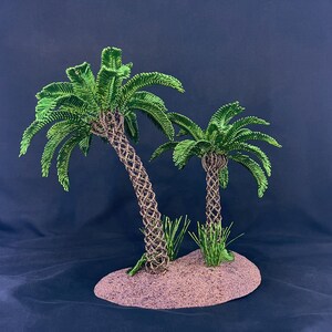 May include: A decorative sculpture featuring two palm trees with intricately woven brown wire trunks and vibrant green wire fronds. The trees are mounted on a textured, oval-shaped base that resembles sandy ground, with small green wire grass accents.