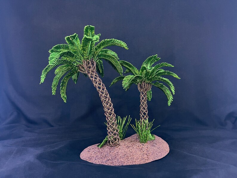 May include: A decorative sculpture featuring two palm trees with intricately woven brown wire trunks and vibrant green wire fronds. The trees are mounted on a textured, oval-shaped base that resembles sandy ground, with small green wire grass accents.
