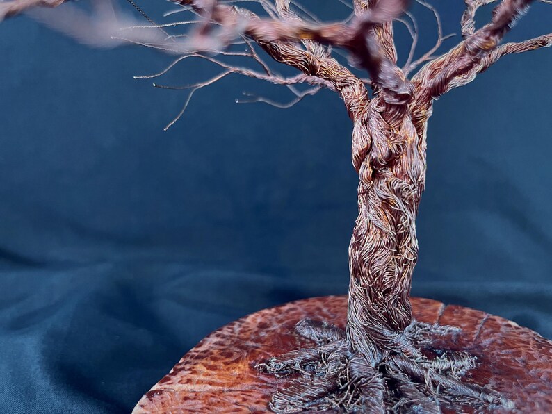 May include: A decorative sculpture of a tree crafted from twisted copper wire. The trunk and branches are formed by numerous thin, metallic strands. The tree is mounted on a circular, polished wooden base with a visible grain and textured surface.