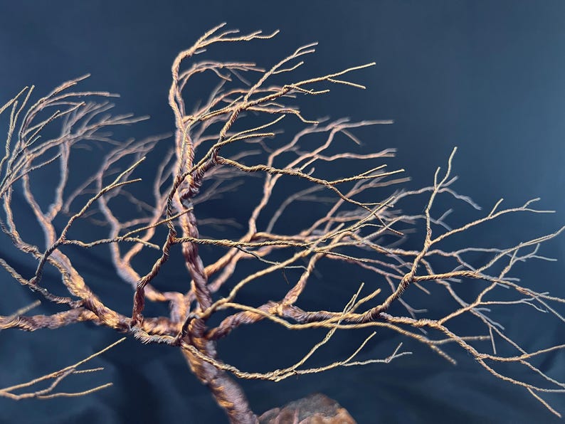 Puede incluir: Una escultura de un &aacute;rbol desnudo hecha de alambre de cobre trenzado. La estructura presenta un tronco grueso y trenzado que se ramifica en numerosas ramitas finas y delicadas que se extienden en varias direcciones, creando una silueta org&aacute;nica compleja.