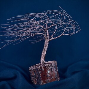 Puede incluir: Una escultura de &aacute;rbol hecha de alambre met&aacute;lico fino de color cobre, con un tronco retorcido y ramas que se extienden horizontalmente hacia un lado. El &aacute;rbol est&aacute; anclado en una base cil&iacute;ndrica de madera natural texturizada de color marr&oacute;n.