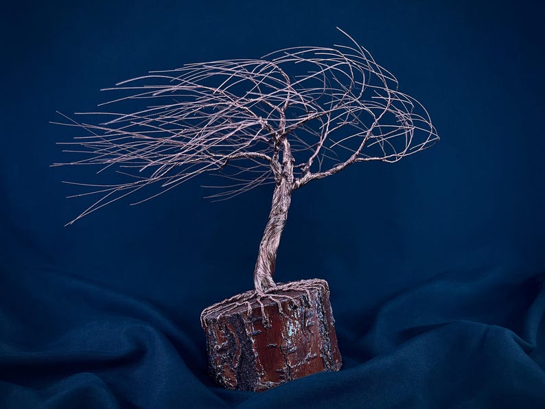 Puede incluir: Una escultura de &aacute;rbol hecha de alambre met&aacute;lico fino de color cobre, con un tronco retorcido y ramas que se extienden horizontalmente hacia un lado. El &aacute;rbol est&aacute; anclado en una base cil&iacute;ndrica de madera natural texturizada de color marr&oacute;n.