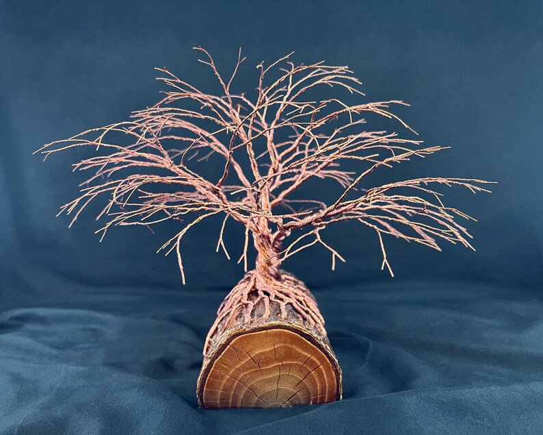 Puede incluir: Una escultura decorativa que presenta un &aacute;rbol de alambre de cobre con ramas finas e intrincadas que se extienden hacia afuera. La base del &aacute;rbol est&aacute; formada por ra&iacute;ces de alambre de cobre envueltas alrededor de un cilindro de madera natural.