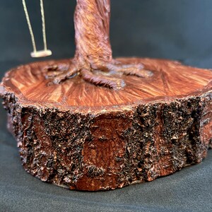 May include: A decorative sculpture featuring a brown wire tree with a textured trunk and roots anchored to a thick, circular wooden base with rough, dark bark edges. A small, light-coloured swing hangs from one of the wire branches of the tree.