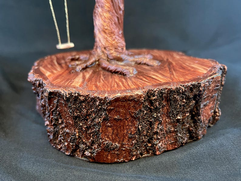 May include: A decorative sculpture featuring a brown wire tree with a textured trunk and roots anchored to a thick, circular wooden base with rough, dark bark edges. A small, light-coloured swing hangs from one of the wire branches of the tree.