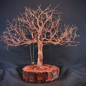 May include: A wire sculpture of a leafless tree with a twisted trunk and intricate branches, mounted on a circular wooden base. A small, thin swing hangs from one of the lower branches. The sculpture features a copper-toned metallic finish.
