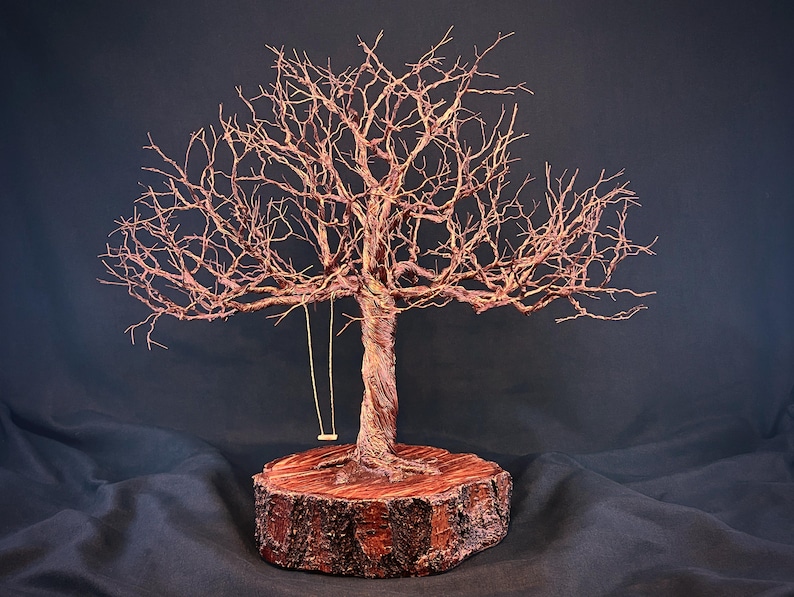 May include: A wire sculpture of a leafless tree with a twisted trunk and intricate branches, mounted on a circular wooden base. A small, thin swing hangs from one of the lower branches. The sculpture features a copper-toned metallic finish.