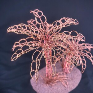 May include: A sculpture of two palm trees crafted from twisted copper wire. The trees feature intricate, open-loop wire foliage and textured trunks, all mounted on a solid, rounded base that resembles a small patch of sandy ground.