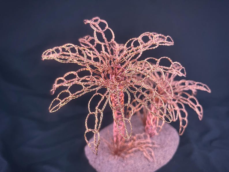 May include: A sculpture of two palm trees crafted from twisted copper wire. The trees feature intricate, open-loop wire foliage and textured trunks, all mounted on a solid, rounded base that resembles a small patch of sandy ground.