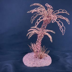 May include: A sculpture of a palm tree crafted from thin copper-colored wire, featuring a woven trunk and detailed fronds. The tree is anchored to a circular, textured base covered in fine, light-colored sand with small wire grass accents.