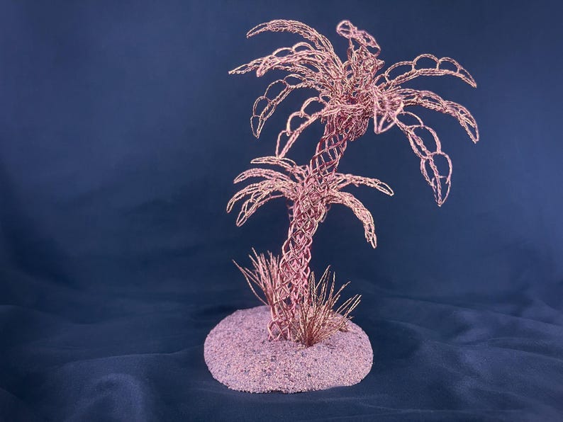 May include: A sculpture of a palm tree crafted from thin copper-colored wire, featuring a woven trunk and detailed fronds. The tree is anchored to a circular, textured base covered in fine, light-colored sand with small wire grass accents.