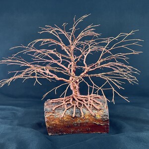 Puede incluir: Una escultura de &aacute;rbol hecha de alambre de cobre fino, con ramas intrincadas y un tronco que se extiende en ra&iacute;ces visibles. Las ra&iacute;ces est&aacute;n ancladas en una pieza rectangular de madera natural con una superficie de corteza rugosa.
