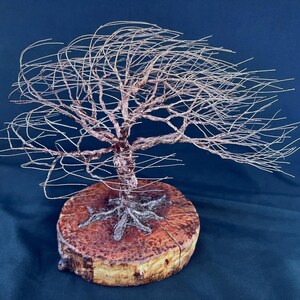 May include: A wire sculpture of a tree with thin, metallic branches extending outward in a wind-swept formation. The tree is anchored to a circular, natural wood slice base with visible bark edges and a textured, reddish-brown grain pattern.