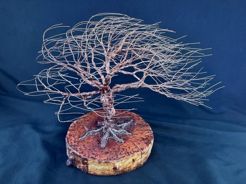 May include: A wire sculpture of a tree with thin, metallic branches extending outward in a wind-swept formation. The tree is anchored to a circular, natural wood slice base with visible bark edges and a textured, reddish-brown grain pattern.