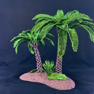 May include: A decorative sculpture featuring two palm trees with vibrant green wire-wrapped fronds and textured brown trunks, set upon a small, oval-shaped brown base with a small green plant accent at the base of the taller tree.