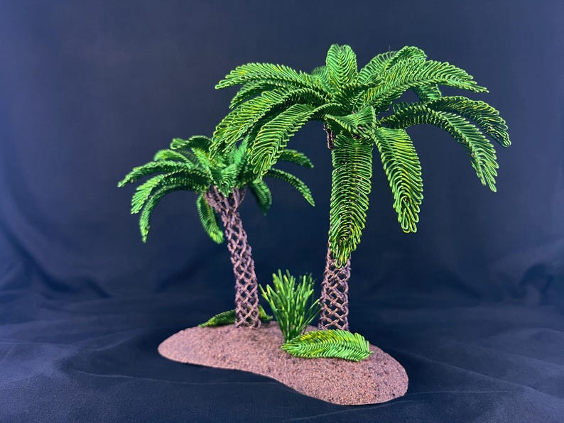 May include: A decorative sculpture featuring two palm trees with vibrant green wire-wrapped fronds and textured brown trunks, set upon a small, oval-shaped brown base with a small green plant accent at the base of the taller tree.