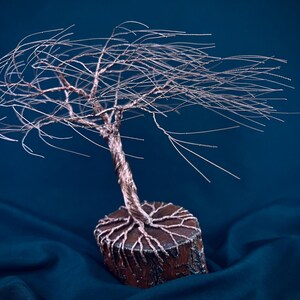 Puede incluir: Una escultura de alambre de un &aacute;rbol con un tronco retorcido de color cobre y ramas finas y extendidas. La base es una rodaja de madera circular con ra&iacute;ces de alambre de cobre que se extienden por la superficie superior con textura.
