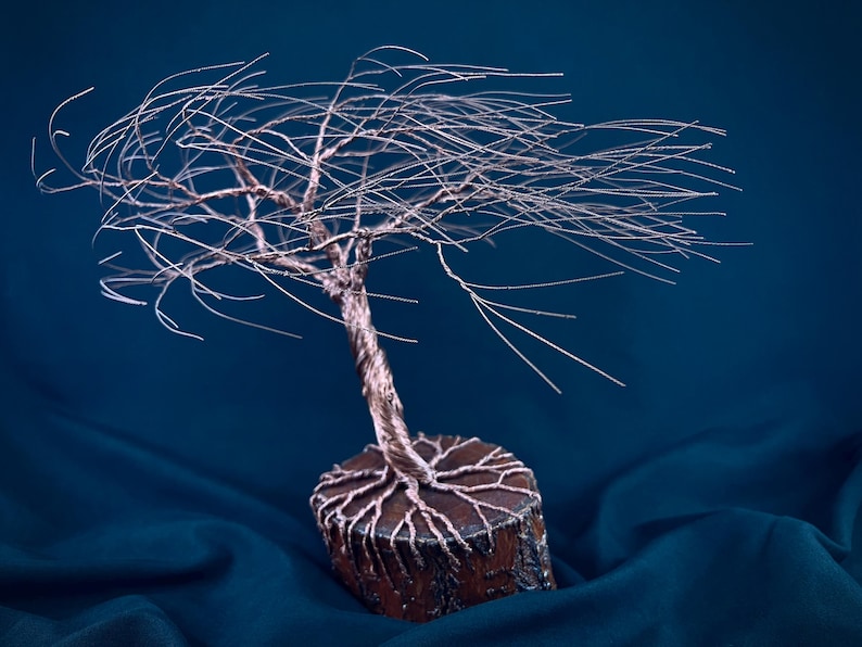 Puede incluir: Una escultura de alambre de un &aacute;rbol con un tronco retorcido de color cobre y ramas finas y extendidas. La base es una rodaja de madera circular con ra&iacute;ces de alambre de cobre que se extienden por la superficie superior con textura.