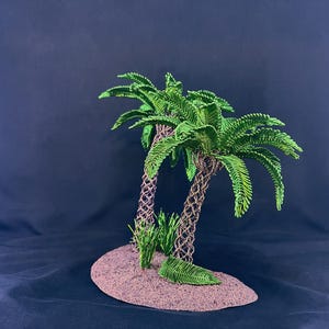 May include: A decorative sculpture featuring two palm trees with textured brown trunks and vibrant green fronds, positioned on a small, oval-shaped brown base with small green grass-like tufts at the base of the trunks.