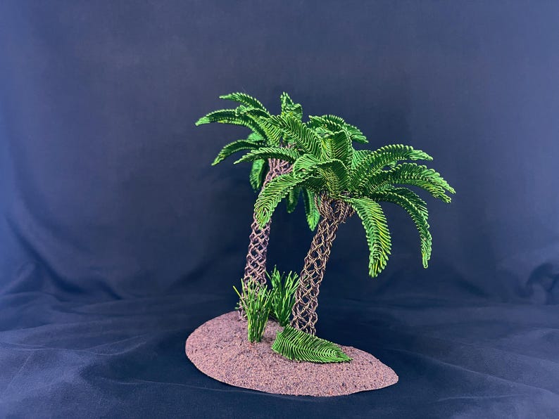 May include: A decorative sculpture featuring two palm trees with textured brown trunks and vibrant green fronds, positioned on a small, oval-shaped brown base with small green grass-like tufts at the base of the trunks.