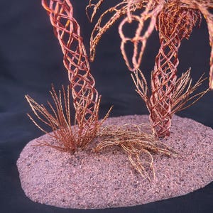May include: A sculpture featuring two palm trees crafted from twisted copper wire, with trunks formed by an intricate lattice pattern and fronds made of thin wire strands, all mounted on a textured, oval-shaped base with a granular surface.
