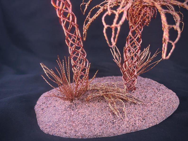 May include: A sculpture featuring two palm trees crafted from twisted copper wire, with trunks formed by an intricate lattice pattern and fronds made of thin wire strands, all mounted on a textured, oval-shaped base with a granular surface.