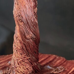 May include: A sculpture of a tree trunk crafted from numerous thin, intertwined copper wires, featuring a textured, fibrous appearance. To the left, a small, light-coloured wooden swing hangs from a single thin wire attached to the copper structure.