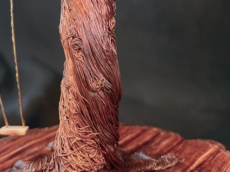 May include: A sculpture of a tree trunk crafted from numerous thin, intertwined copper wires, featuring a textured, fibrous appearance. To the left, a small, light-coloured wooden swing hangs from a single thin wire attached to the copper structure.