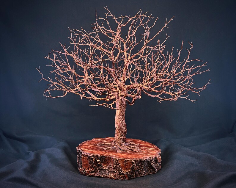 May include: A copper wire sculpture of a leafless tree with a twisted trunk and intricate, spreading branches. The sculpture is mounted on a circular, reddish-brown wooden base with visible bark texture around the outer edge of the wood.