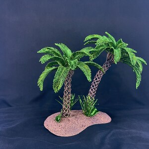 May include: Two palm trees crafted from twisted brown wire trunks and vibrant green wire fronds, standing on a textured, sandy-colored base. Small patches of green wire grass are positioned at the base of each tree trunk on the oval-shaped platform.