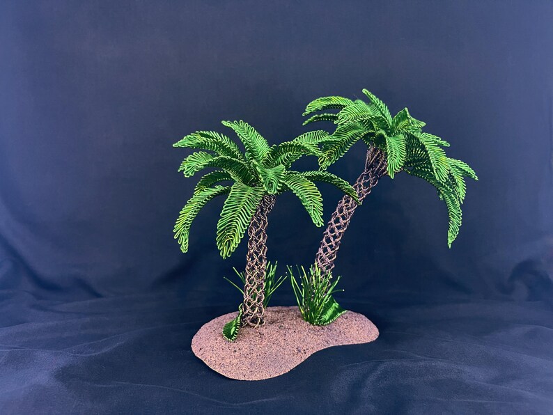 May include: Two palm trees crafted from twisted brown wire trunks and vibrant green wire fronds, standing on a textured, sandy-colored base. Small patches of green wire grass are positioned at the base of each tree trunk on the oval-shaped platform.