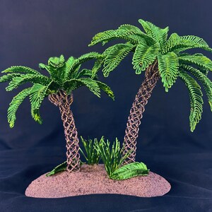 May include: Two palm trees crafted from green wire with textured brown wire trunks, standing on a small, rounded, sandy-colored base. The palm fronds are made of looped green wire, and small green wire grass accents are at the base of the trees.