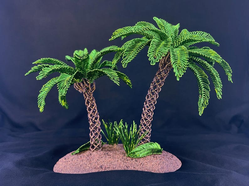 May include: Two palm trees crafted from green wire with textured brown wire trunks, standing on a small, rounded, sandy-colored base. The palm fronds are made of looped green wire, and small green wire grass accents are at the base of the trees.