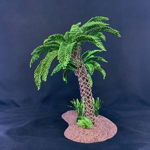 May include: A wire sculpture of a palm tree featuring a brown woven trunk and vibrant green wire fronds. The tree is mounted on a textured, sandy-colored base with small green wire grass accents at the bottom of the trunk.