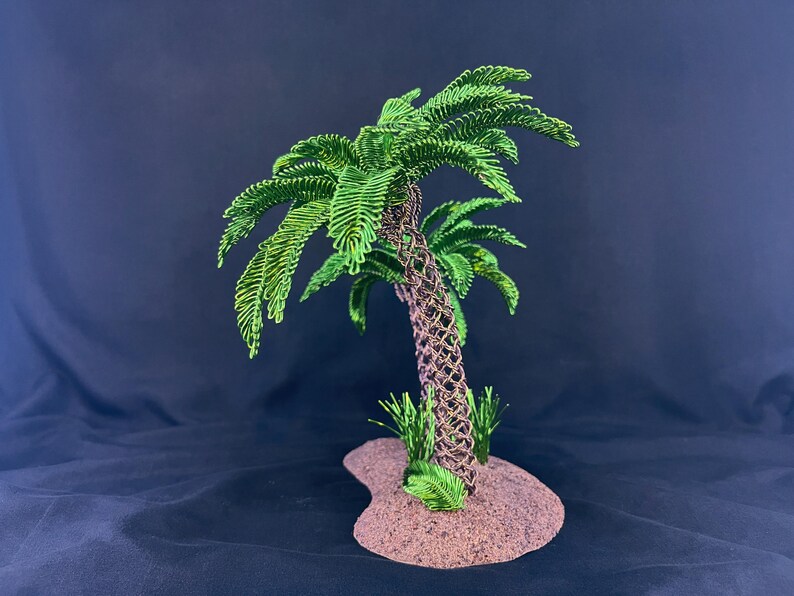 May include: A wire sculpture of a palm tree featuring a brown woven trunk and vibrant green wire fronds. The tree is mounted on a textured, sandy-colored base with small green wire grass accents at the bottom of the trunk.