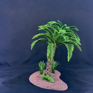 May include: A decorative sculpture of a palm tree crafted from green wire, featuring a textured brown wire trunk. The tree is mounted on a small, oval-shaped base with a sandy, tan-colored surface, accompanied by two small green wire grass tufts.