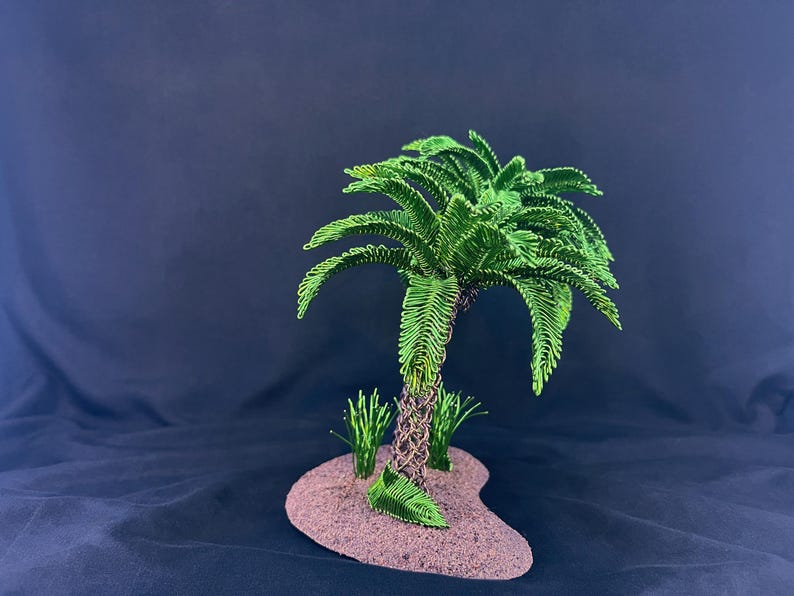 May include: A decorative sculpture of a palm tree crafted from green wire, featuring a textured brown wire trunk. The tree is mounted on a small, oval-shaped base with a sandy, tan-colored surface, accompanied by two small green wire grass tufts.