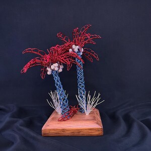 May include: A decorative sculpture featuring two palm trees crafted from twisted wire. The trunks are blue, the fronds are red, and clusters of white beads represent coconuts. The trees are mounted on a square wooden base with white wire accents.