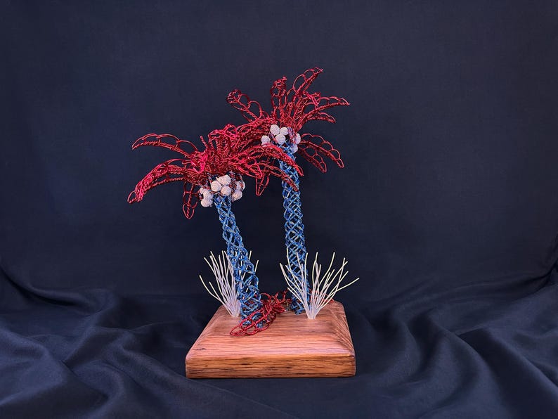 May include: A decorative sculpture featuring two palm trees crafted from twisted wire. The trunks are blue, the fronds are red, and clusters of white beads represent coconuts. The trees are mounted on a square wooden base with white wire accents.