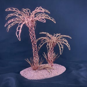 May include: A decorative sculpture featuring two palm trees crafted from copper-colored wire. The trees have intricate, woven trunks and delicate, spreading fronds. They are mounted on a textured, oval-shaped base that is light pink in color.