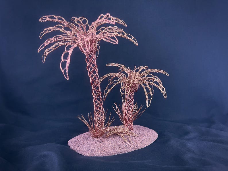 May include: A decorative sculpture featuring two palm trees crafted from copper-colored wire. The trees have intricate, woven trunks and delicate, spreading fronds. They are mounted on a textured, oval-shaped base that is light pink in color.