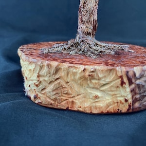 May include: A sculpture featuring a metallic wire tree trunk with visible roots anchored onto a circular, light-colored wooden base. The base has a textured, carved exterior surface with natural wood grain patterns and a smooth, flat top surface.