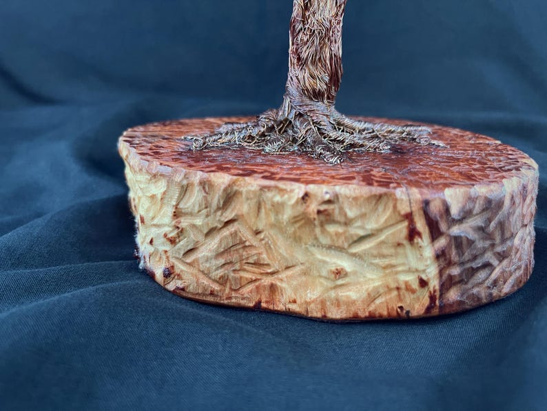 May include: A sculpture featuring a metallic wire tree trunk with visible roots anchored onto a circular, light-colored wooden base. The base has a textured, carved exterior surface with natural wood grain patterns and a smooth, flat top surface.