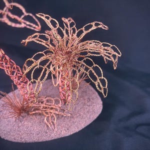 May include: A sculpture of a palm tree crafted from thin copper wire, featuring intricate woven fronds and a textured trunk. The structure is mounted on a small, oval-shaped base with a granular, sandy-colored surface and additional wire foliage.