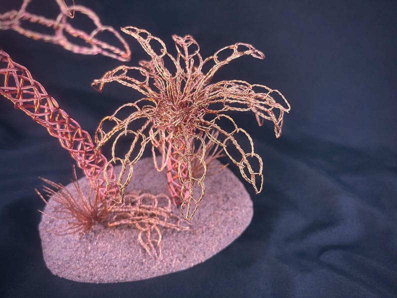 May include: A sculpture of a palm tree crafted from thin copper wire, featuring intricate woven fronds and a textured trunk. The structure is mounted on a small, oval-shaped base with a granular, sandy-colored surface and additional wire foliage.