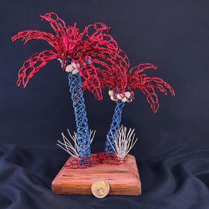 May include: A decorative sculpture featuring two palm trees crafted from red and blue wire, mounted on a rectangular wooden base. The trunks are woven blue wire, the fronds are red wire, and small white shells are attached. A US quarter coin sits at the base.