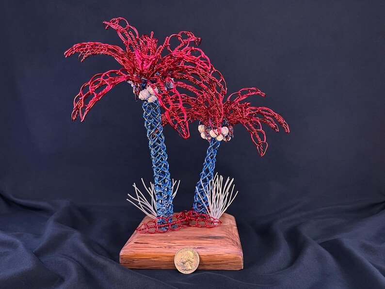 May include: A decorative sculpture featuring two palm trees crafted from red and blue wire, mounted on a rectangular wooden base. The trunks are woven blue wire, the fronds are red wire, and small white shells are attached. A US quarter coin sits at the base.