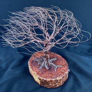 May include: A sculpture of a tree crafted from thin, metallic copper-colored wire, featuring a wide, spreading canopy of branches. The wire trunk and exposed roots are anchored securely onto a circular, polished wooden base with a textured grain.