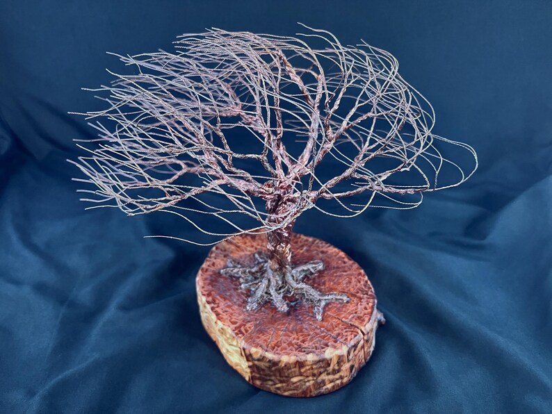 May include: A sculpture of a tree crafted from thin, metallic copper-colored wire, featuring a wide, spreading canopy of branches. The wire trunk and exposed roots are anchored securely onto a circular, polished wooden base with a textured grain.