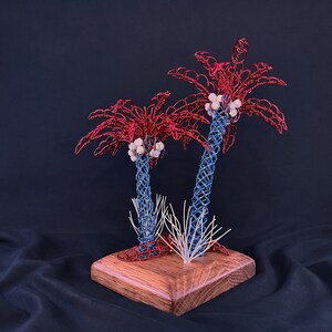 May include: A decorative sculpture featuring two palm trees crafted from twisted wire. The trunks are blue, the fronds are red, and the coconuts are white. The trees are mounted on a square wooden base with a small cluster of white wire grass.
